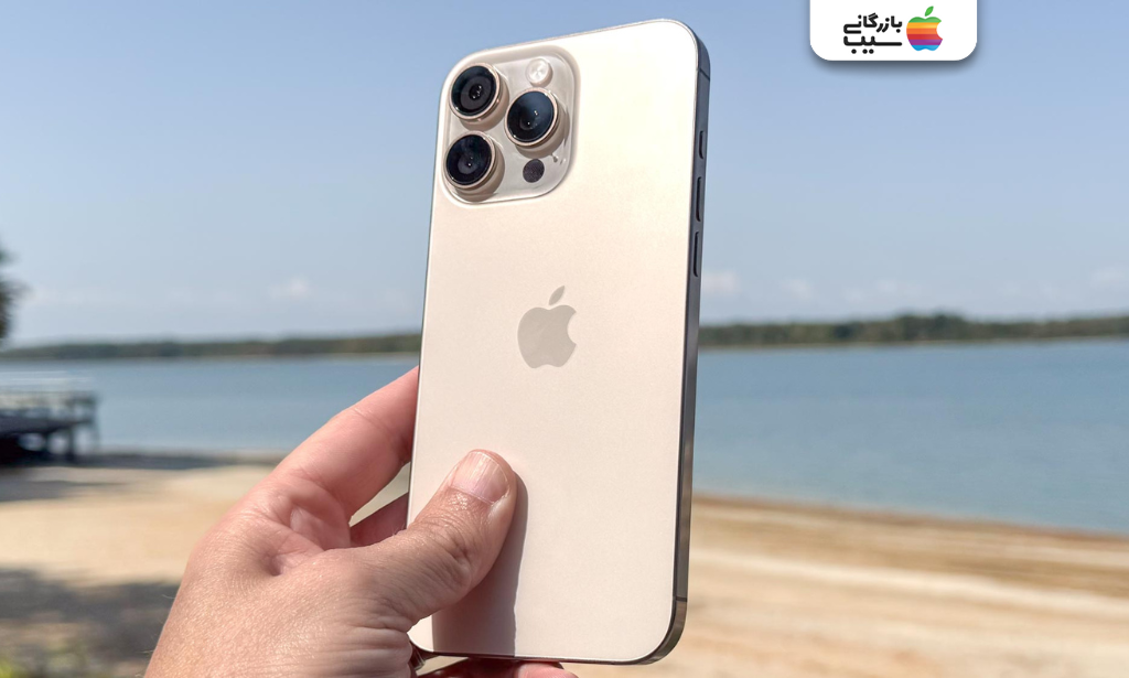 نمای پشت آیفون طلایی سه دوربین در دست کاربر کنار ساحل و آسمان آبی